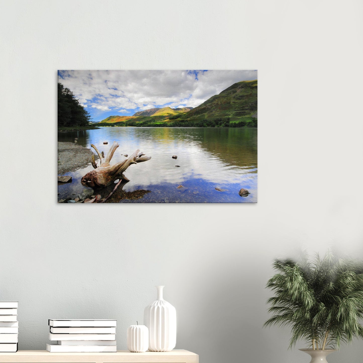 Canvas print showing the tranquil beauty of Buttermere Lake with driftwood foreground, mirrored sky, and lush green fells under a partly cloudy sky.