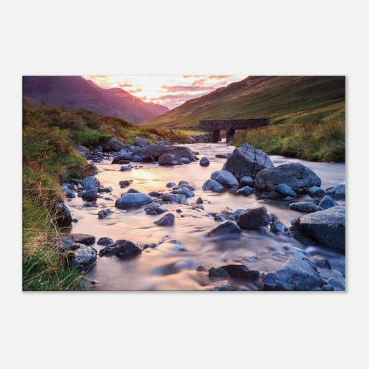 Canvas art of Honister Pass at dusk with warm hues, a silky ribbon of water, and a heritage stone bridge nestled among stones.