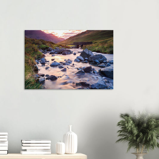 Canvas print of Honister Pass in the Lake District at golden hour with a winding stream and stone bridge in warm light.