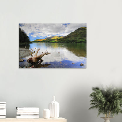 Canvas print showing the tranquil beauty of Buttermere Lake with driftwood foreground, mirrored sky, and lush green fells under a partly cloudy sky.