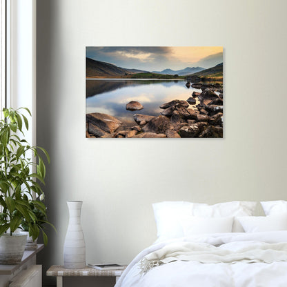 Softly lit sky and tranquil lake waters with foreground rocks guiding eyes toward Snowdonia peaks
